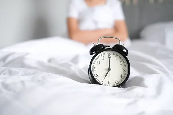 An alarm clock on a bed with a woman in the background